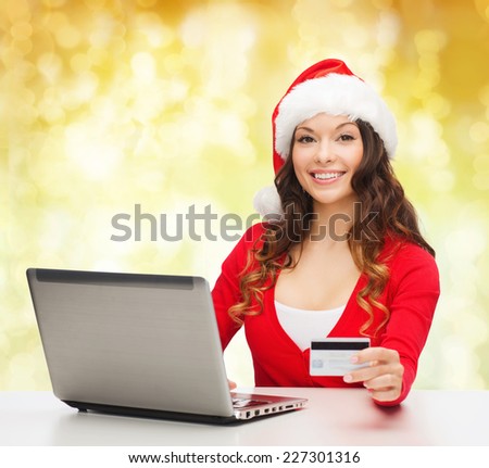 christmas, holidays, technology and shopping concept - smiling woman in santa helper hat with credit card and laptop computer over yellow lights background