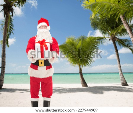 christmas, holidays and people concept - man in costume of santa claus with gift box over tropical beach background