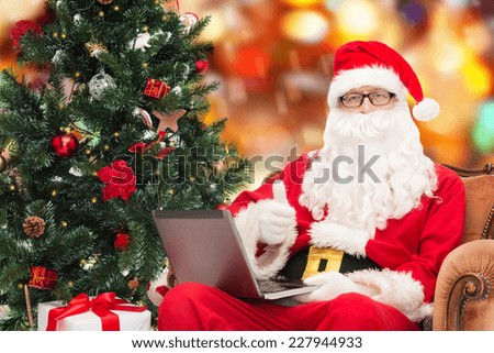 technology, holidays and people concept - man in costume of santa claus with laptop computer, gifts and christmas tree sitting in armchair over red lights background
