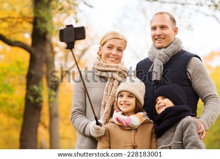 family, childhood, season, technology and people concept - happy family photographing with smartphone and selfie stick in autumn park