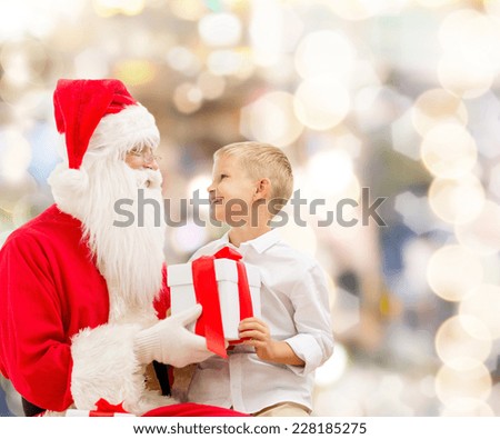 holidays, christmas, childhood and people concept - smiling little boy with santa claus and gifts over lights background