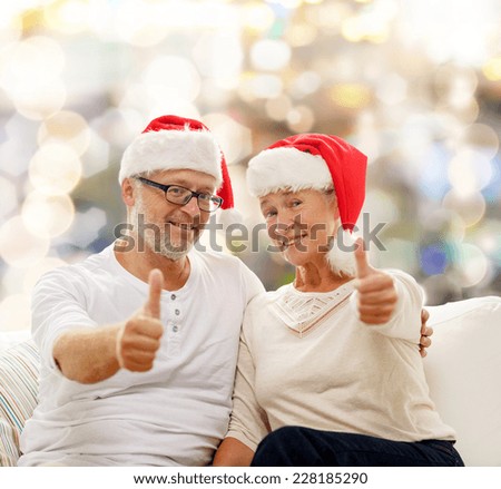 family, holidays, christmas, age and people concept - happy senior couple in santa helper hats sitting on sofa over lights background and showing thumbs up gesture