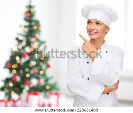 cooking, holidays and people concept - smiling female chef, cook or baker pointing finger up over living room and christmas tree background