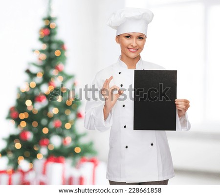 cooking, advertisement, holidays and people concept - smiling female chef, cook or baker with blank black menu paper showing ok gesture over living room and christmas tree background