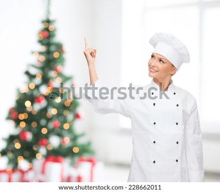 cooking, advertisement, holidays and people concept - smiling female chef, cook or baker pointing finger up to something over living room and christmas tree background