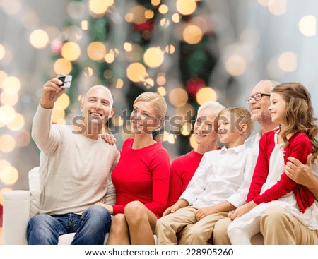 family, holidays, generation, christmas and people concept - smiling family with camera taking selfie and sitting on couch over tree lights background