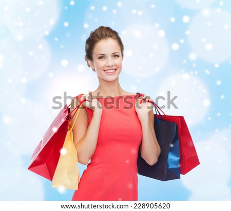 shopping, sale, christmas, holidays and people concept - smiling elegant woman in red dress with shopping bags over blue lights and snow background
