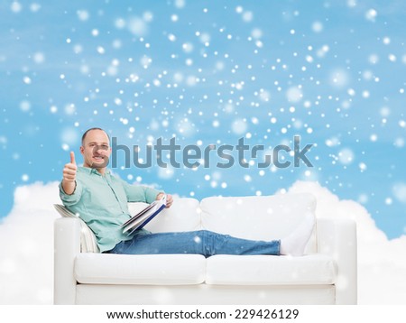 people, leisure and happiness concept - smiling man with book lying on sofa and showing thumbs up over blue sky with cloud and snow background