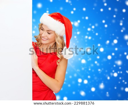 winter holidays, christmas, advertising and people concept - smiling young woman in santa helper hat with white blank billboard over blue snowy background