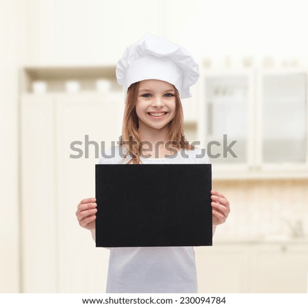 cooking, childhood, advertisement and people concept - smiling little chef girl, cook or baker with blank black paper over kitchen background
