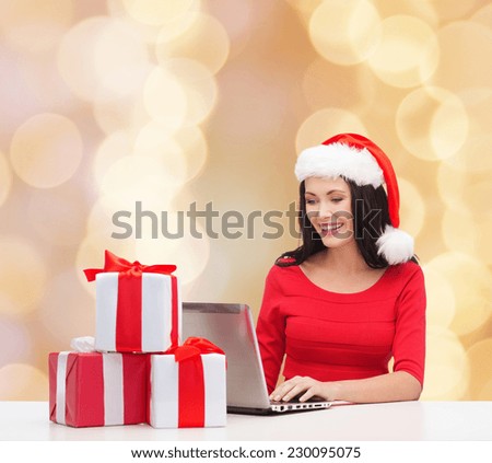 christmas, holidays, technology and people concept - smiling woman in santa helper hat with gift boxes and laptop computer over beige background