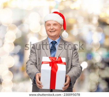 business, christmas, presents and people concept - smiling senior man in suit and santa helper hat with gift over lights background