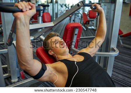 sport, equipment, technology, lifestyle and people concept - young man with earphones flexing chest muscles on gym machine