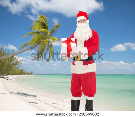 christmas, holidays, travel and people concept - man in costume of santa claus with gift box over tropical beach background