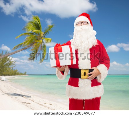 christmas, travel, holidays and people concept - man in costume of santa claus with gift box over tropical beach background