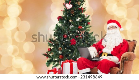 technology, holidays and people concept - man in costume of santa claus with laptop computer, gifts and christmas tree sitting in armchair over beige lights background
