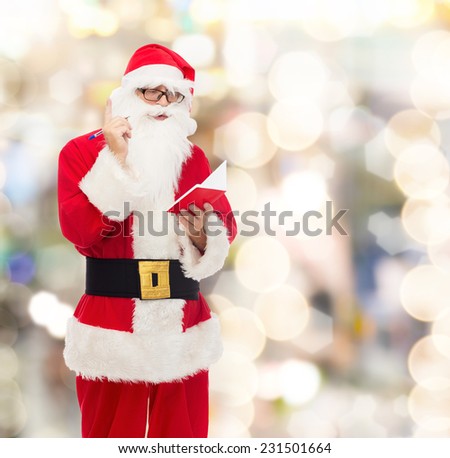 christmas, holidays and people concept - man in costume of santa claus with notepad and pen over lights background