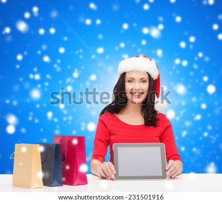 christmas, technology and people concept - smiling woman in santa helper hat with shopping bags and tablet pc computer over blue snowy background