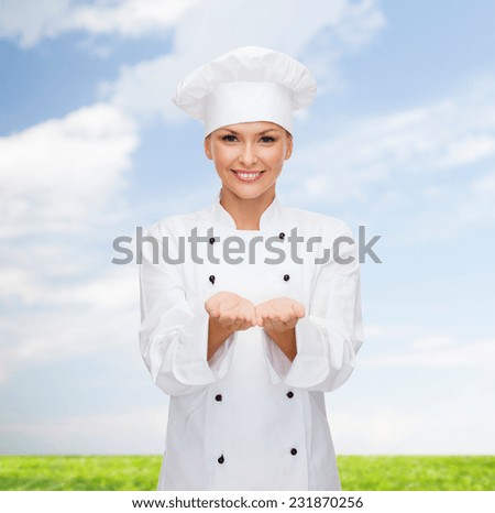 cooking, advertisement and people concept - smiling female chef, cook or baker holding something on palms of hand over blue sky and grass background