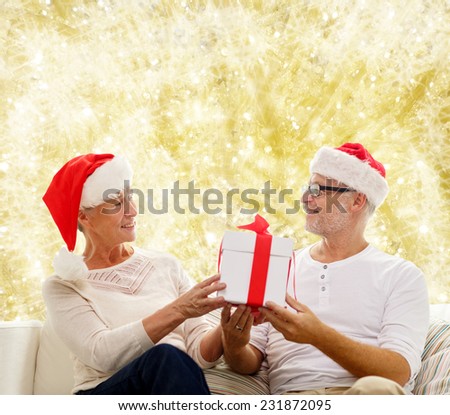 family, holidays, christmas, age and people concept - happy senior couple in santa helper hats with gift box over yellow background