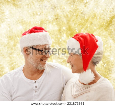family, holidays, christmas, age and people concept - happy senior couple in santa helper hats sitting on sofa over yellow lights background