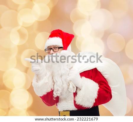 christmas, holidays and people concept - man in costume of santa claus with bag pointing finger over beige lights background