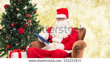 technology, holidays and people concept - man in costume of santa claus with tablet pc computer, gifts and christmas tree sitting in armchair over yellow lights background