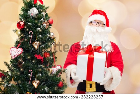 christmas, holidays and people concept - man in costume of santa claus with gift box and tree over beige lights background