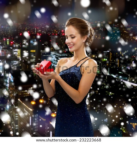 holidays, people and christmas concept - smiling woman in dress holding red gift box over snowy night city background
