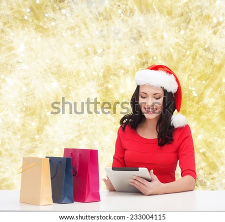 christmas, technology and people concept - smiling woman in santa helper hat with shopping bags and tablet pc computer over yelolow lights background