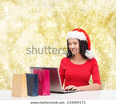 christmas, holidays, technology and people concept - smiling woman in santa helper hat with shopping bags and laptop computer over yellow lights background