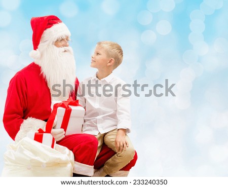 holidays, christmas, childhood and people concept - smiling little boy with santa claus and gifts over blue lights background