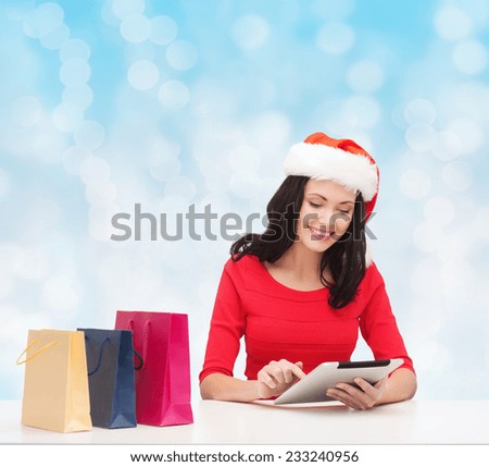christmas, technology and people concept - smiling woman in santa helper hat with shopping bags and tablet pc computer over blue lights background