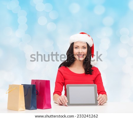 christmas, technology and people concept - smiling woman in santa helper hat with shopping bags and tablet pc computer over blue lights background