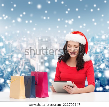 christmas, technology and people concept - smiling woman in santa helper hat with shopping bags and tablet pc computer over snowy city background