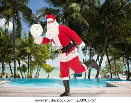 christmas, holidays, travel and people concept - man in costume of santa claus running with clock showing twelve over swimming pool on tropical beach background