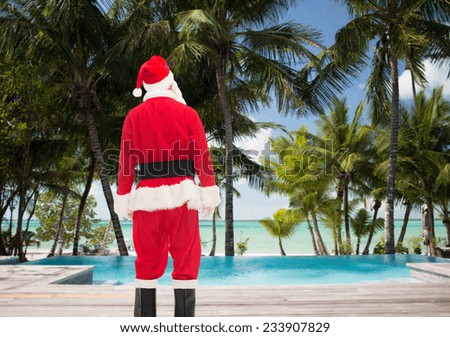 christmas, holidays and people concept - man in costume of santa claus from back over tropical beach and swimming pool background