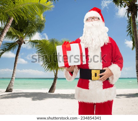christmas, holidays, travel and people concept - man in costume of santa claus with gift box over tropical beach background