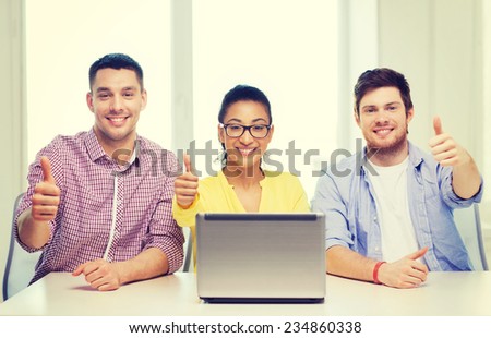 education, technology, business, startup and office concept - three smiling colleagues with laptop in office showing thumbs up