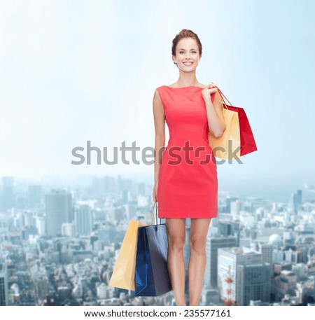 shopping, sale and holidays concept - smiling elegant woman in red dress with shopping bags over city background