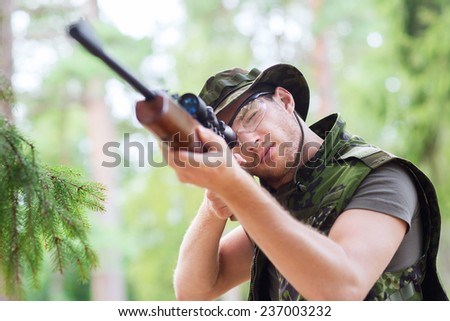hunting, war, army and people concept - young soldier, ranger or hunter with gun aiming and shooting in forest