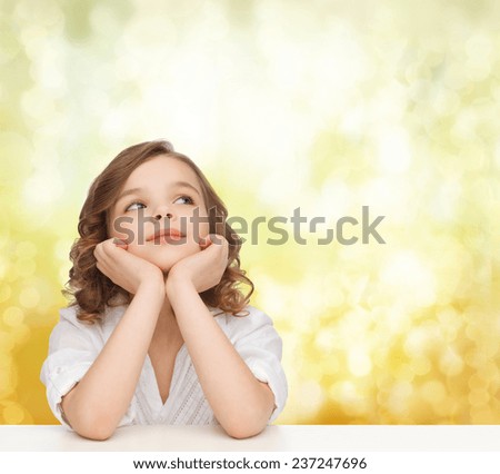 children, childhood, holidays and happy people concept - beautiful girl sitting at table, looking up and dreaming over yellow lights background