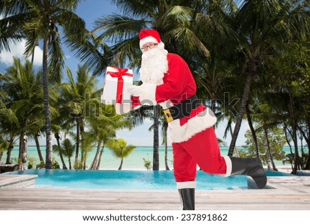 christmas, holidays, travel and people concept - man in costume of santa claus running with gift box over tropical beach and swimming pool background
