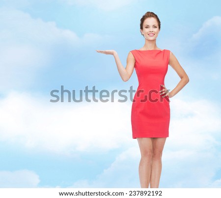 advertising, holidays and people concept - smiling young woman in red dress holding something on palm of her hand over blue cloudy sky background