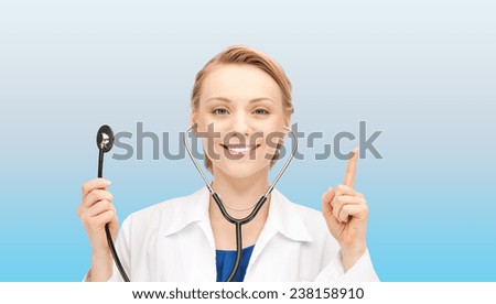 medicine, people, profession and healthcare concept - smiling young female doctor in white coat holding stethoscope and pointing finger up over blue background