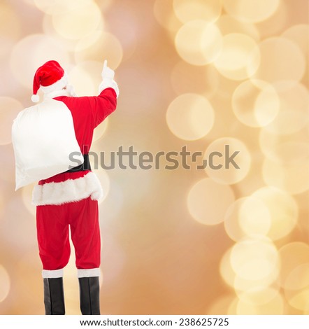 christmas, holidays and people concept - man in costume of santa claus with bag pointing finger from back over yellow lights background over beige lights background