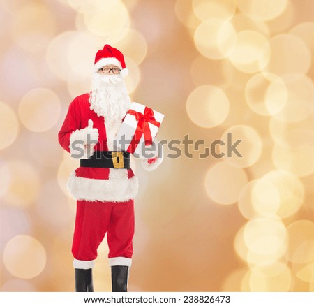 christmas, holidays and people concept - man in costume of santa claus with gift box showing thumbs up gesture over beige lights background