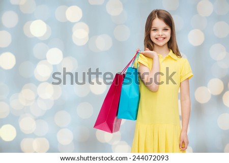 shopping, happiness and people concept - smiling little girl in yellow dress with shopping bags over holidays lights background