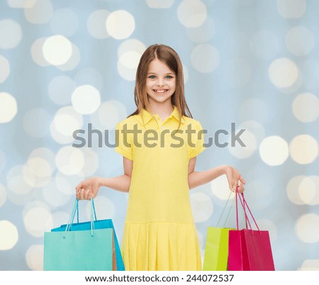 shopping, happiness and people concept - smiling little girl in yellow dress with shopping bags over holidays lights background