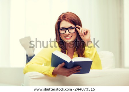 leasure and home concept - smiling teenage girl in eyeglasses reading book and sitting on couch at home